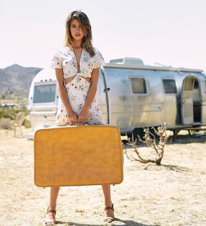 Woman in white floral dress holding yellow suitcase in front of Airstream trailer in desert