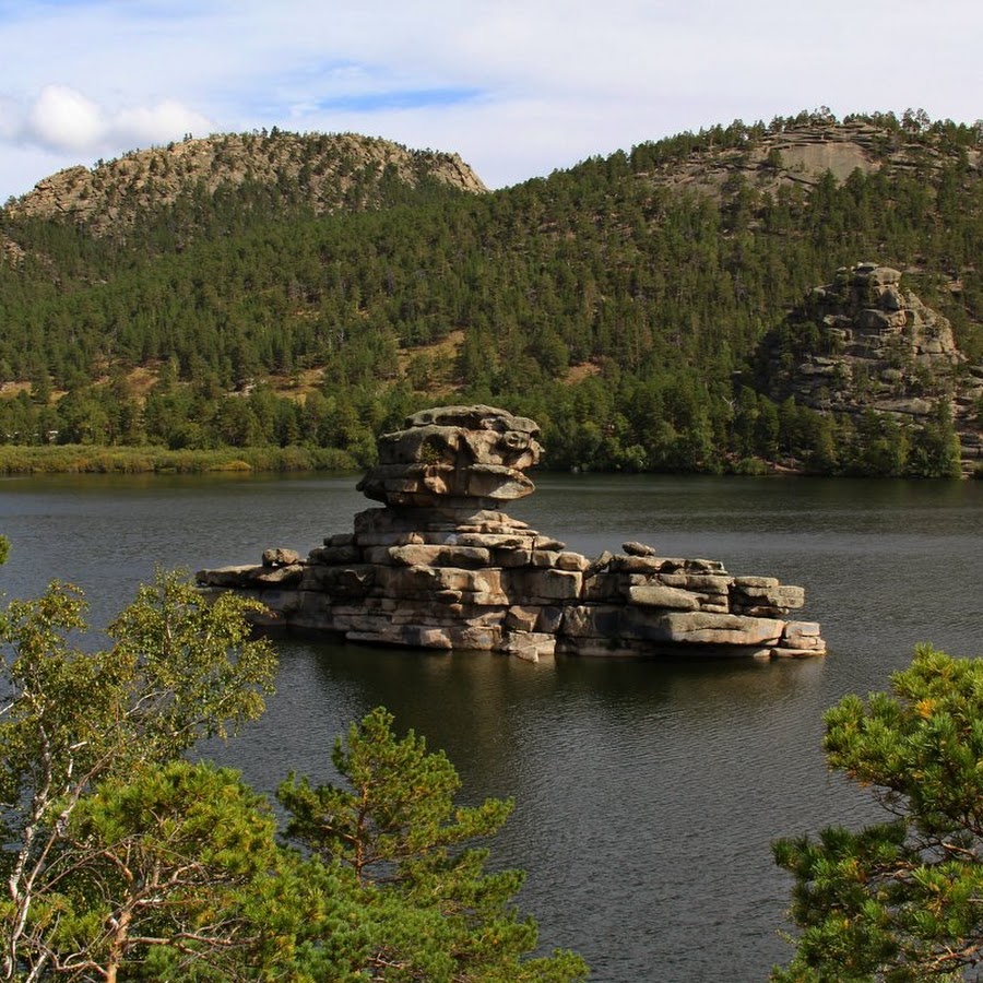 Lake with balanced rock formation on rocky island surrounded by pine-covered hills and granite cliffs