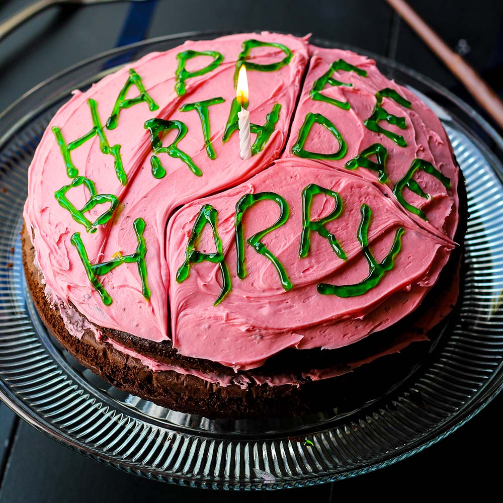 Pink frosted birthday cake with green text reading Happy Birthday Harry on glass cake stand