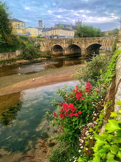 Stone bridge spanning river with vibrant flower gardens along the bank and buildings in background