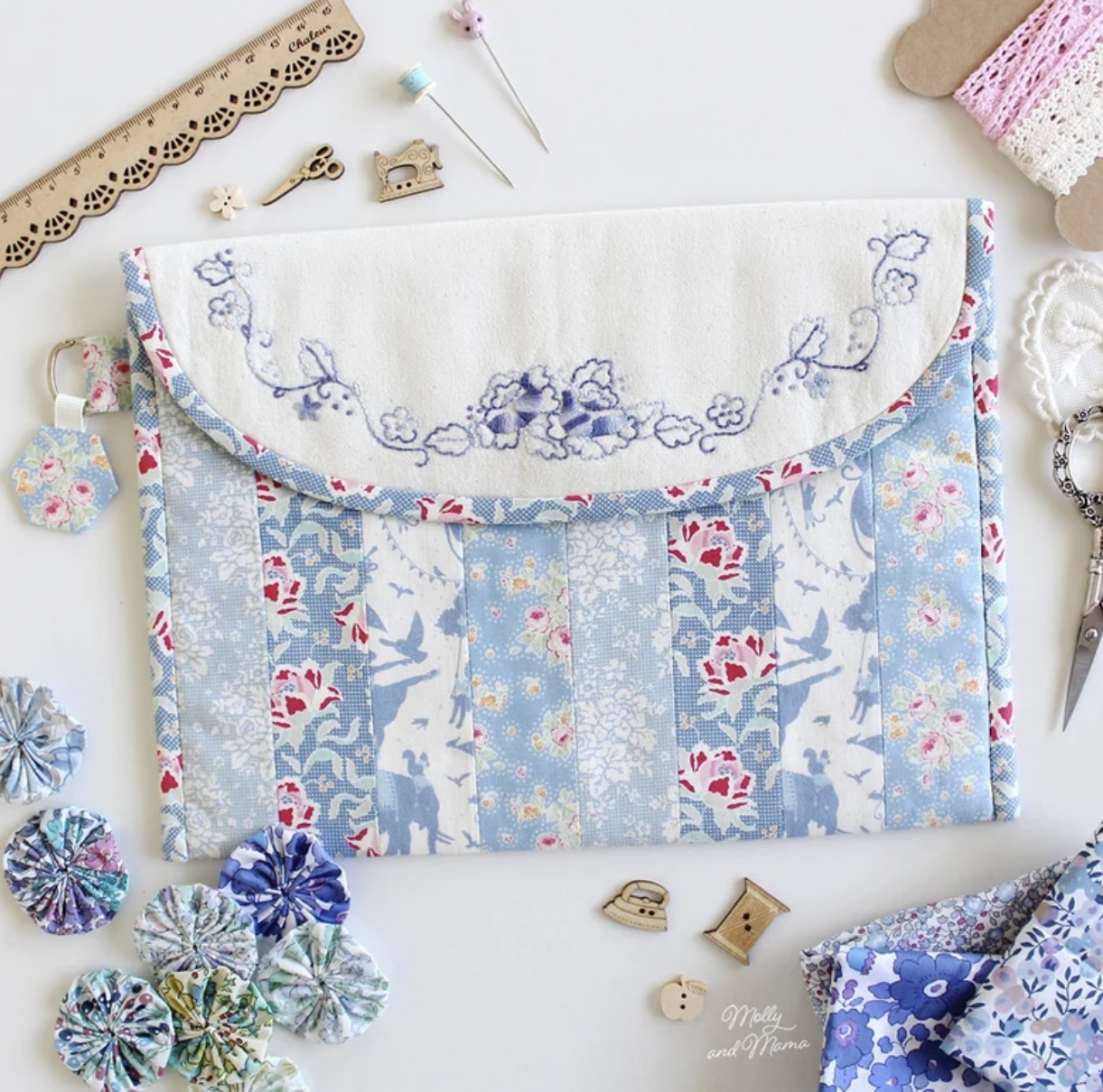 Handmade fabric pouch with embroidered flap and floral patchwork panels, displayed with sewing supplies