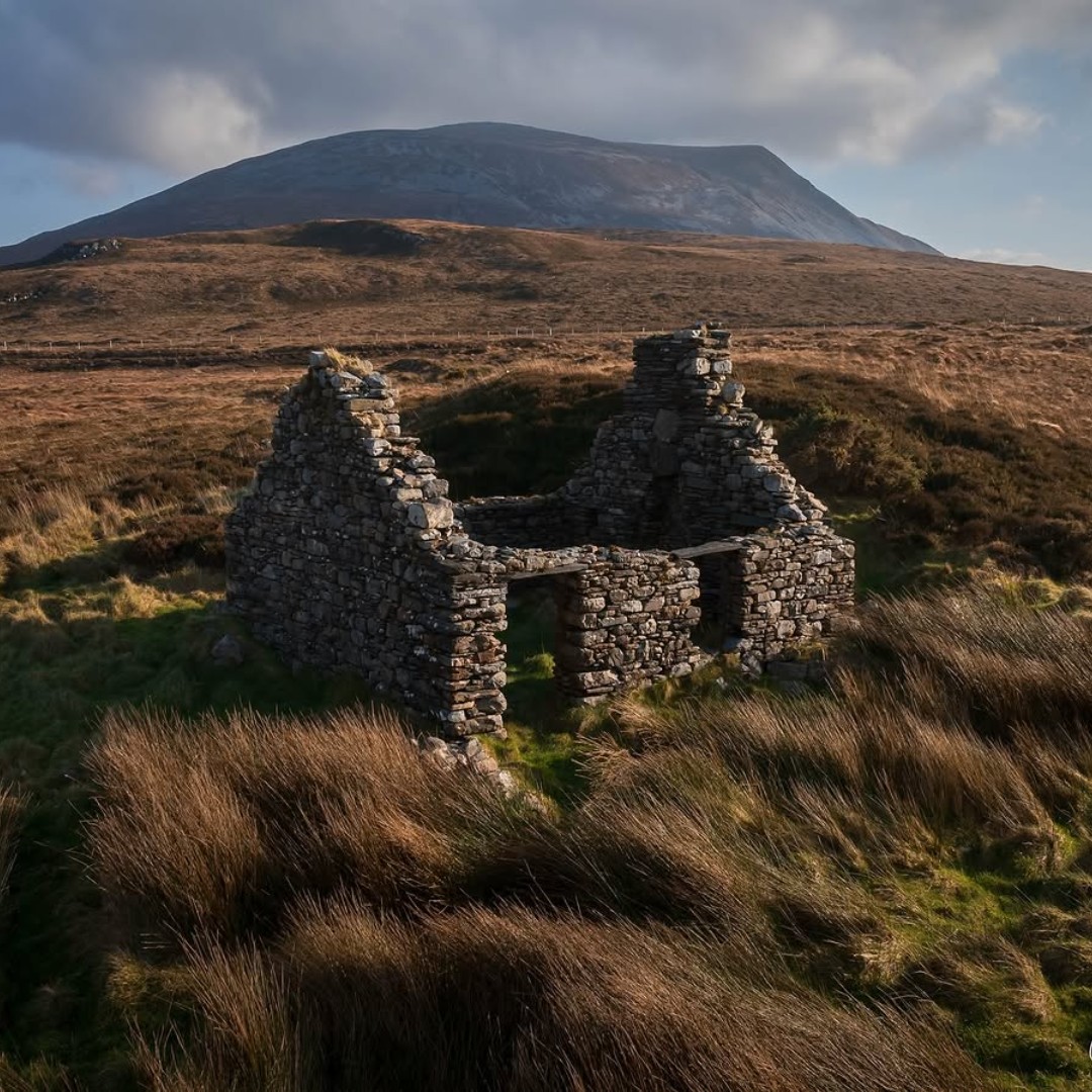 Crumbling stone cottage ruins on grassy moorland beneath a large mountain and cloudy sky