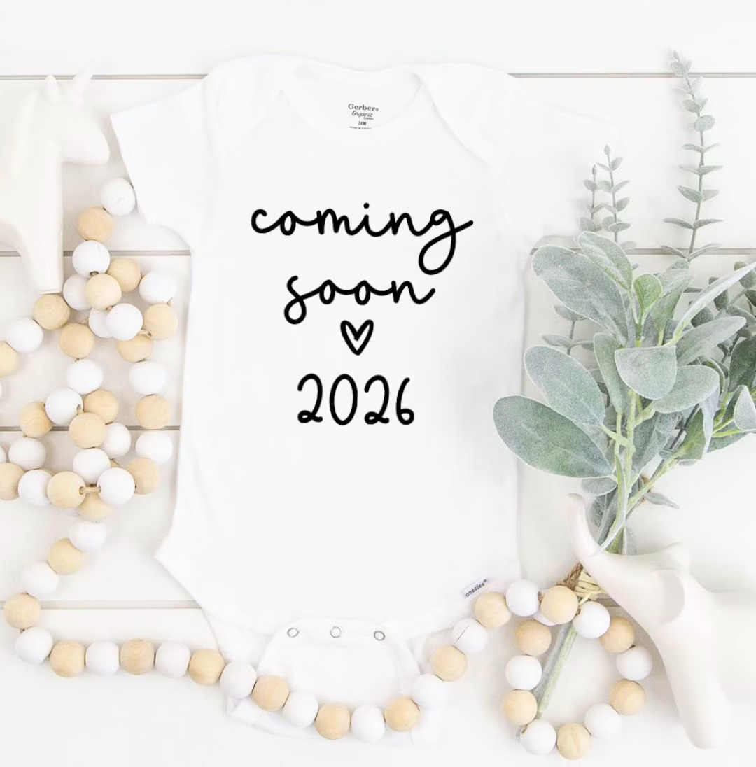 White baby onesie with 'coming soon 2026' text, styled with pom-pom garlands and green leaves