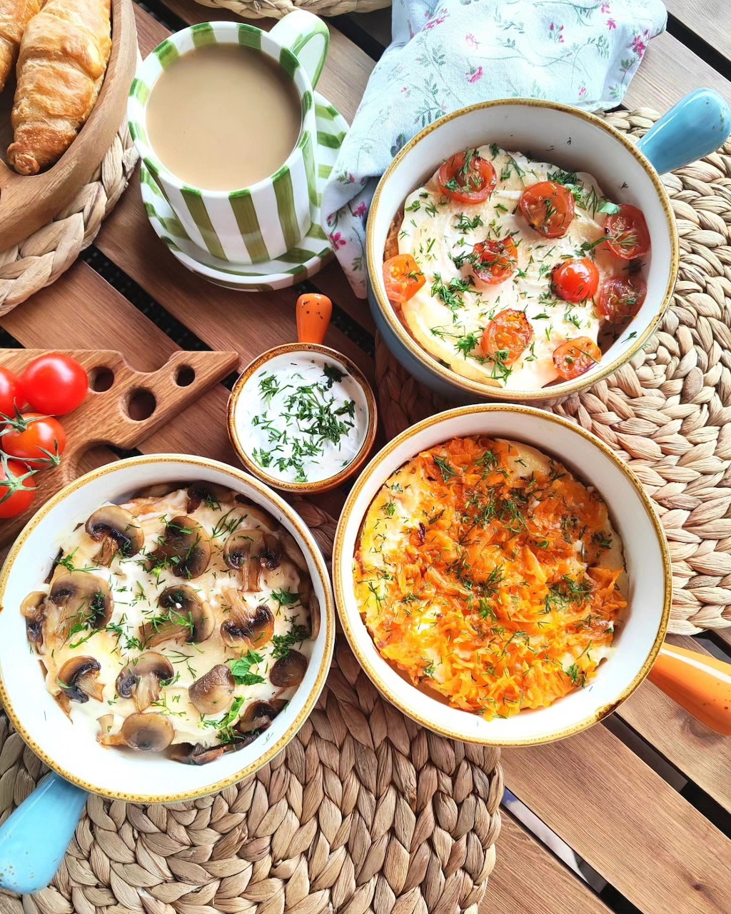 Overhead view of four baked egg ramekins with mushrooms, tomatoes, and cheese, alongside coffee and croissants