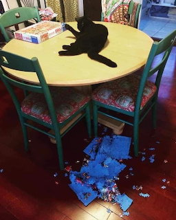 Black cat on round table with jigsaw puzzle pieces scattered on red floor below