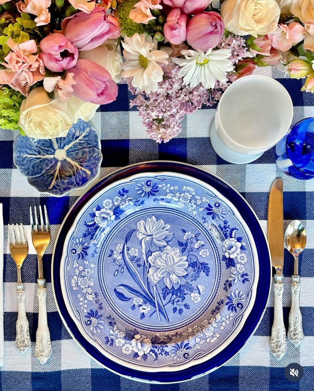 Overhead table setting with blue floral plates, checkered cloth, silverware, cup, and pink flowers