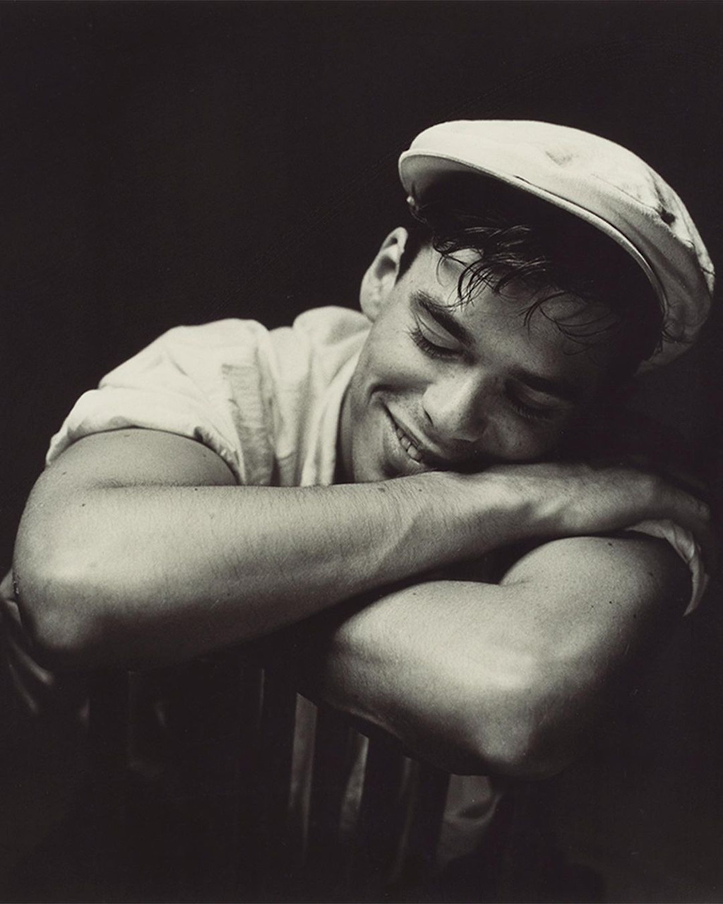 Black and white photo of a smiling young man in a flat cap resting his chin on crossed arms
