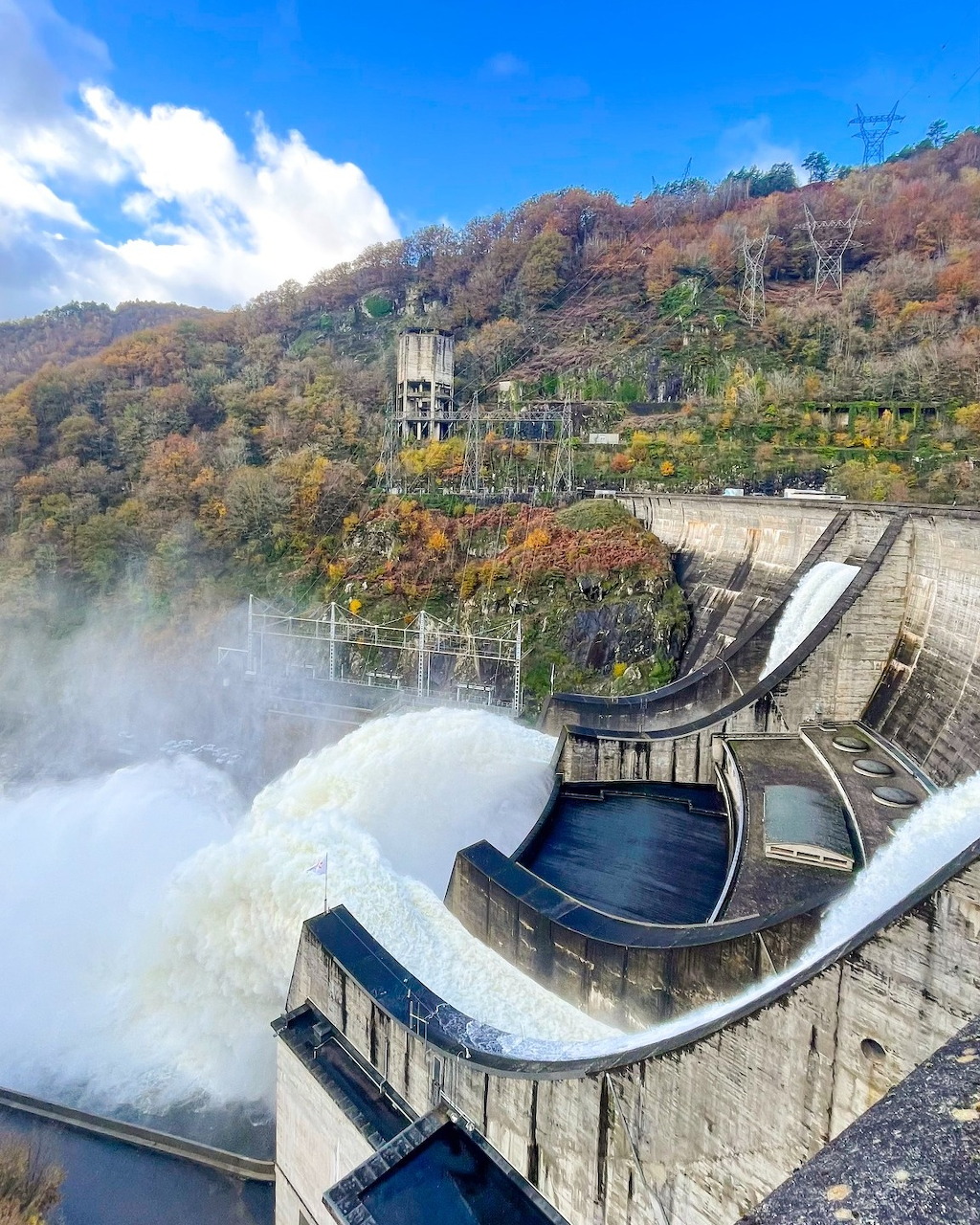Concrete dam discharging water from spillways surrounded by autumn forested hillsides