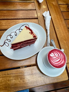Slice of red velvet cake with white frosting next to pink latte with decorative foam art on wooden table