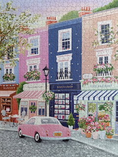 Illustrated village street with pastel buildings, pink van, bookshop, and florist shop with flowers