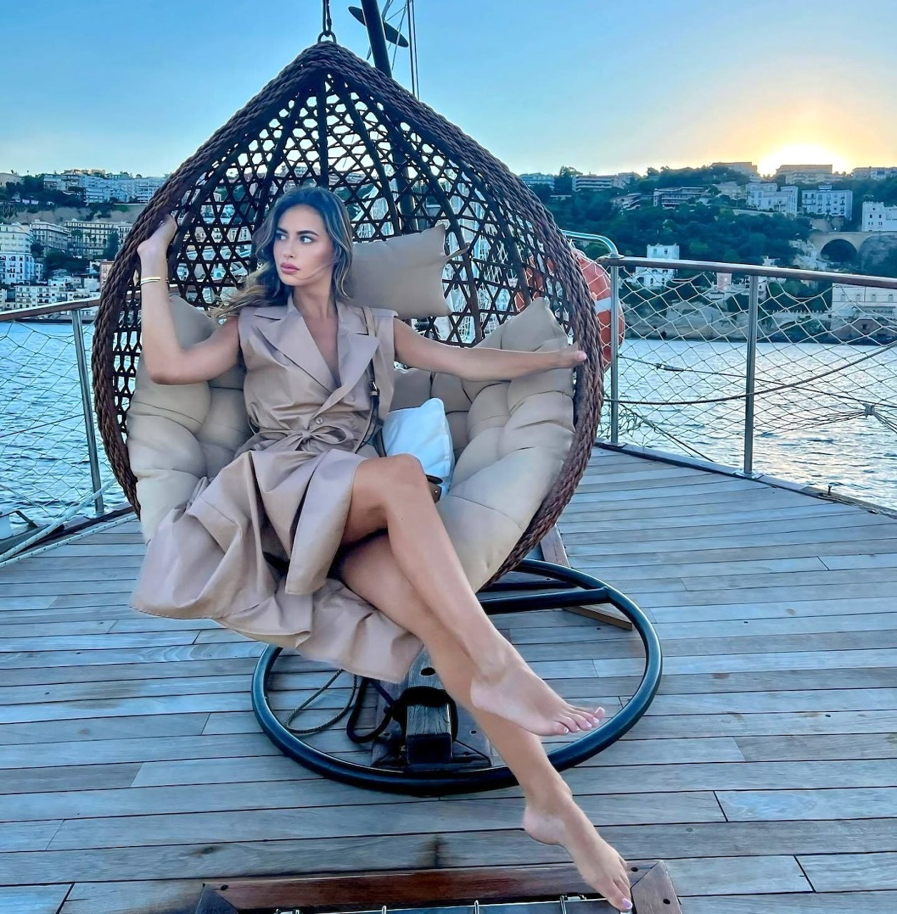 Woman sitting in a wicker hanging egg chair on a deck by a river at sunset