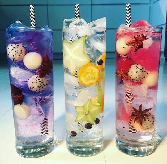 Three tall glasses with colorful layered iced drinks, fruit slices, edible flowers, and striped straws