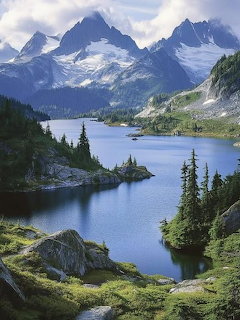 Alpine lake with jagged mountain peaks, evergreen trees, and grassy slopes reflecting in calm blue water