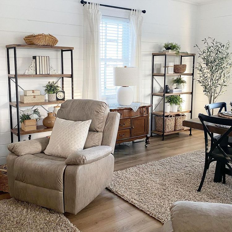 Farmhouse living room with beige armchair, shelving units, area rug, and natural light from window with white curtains