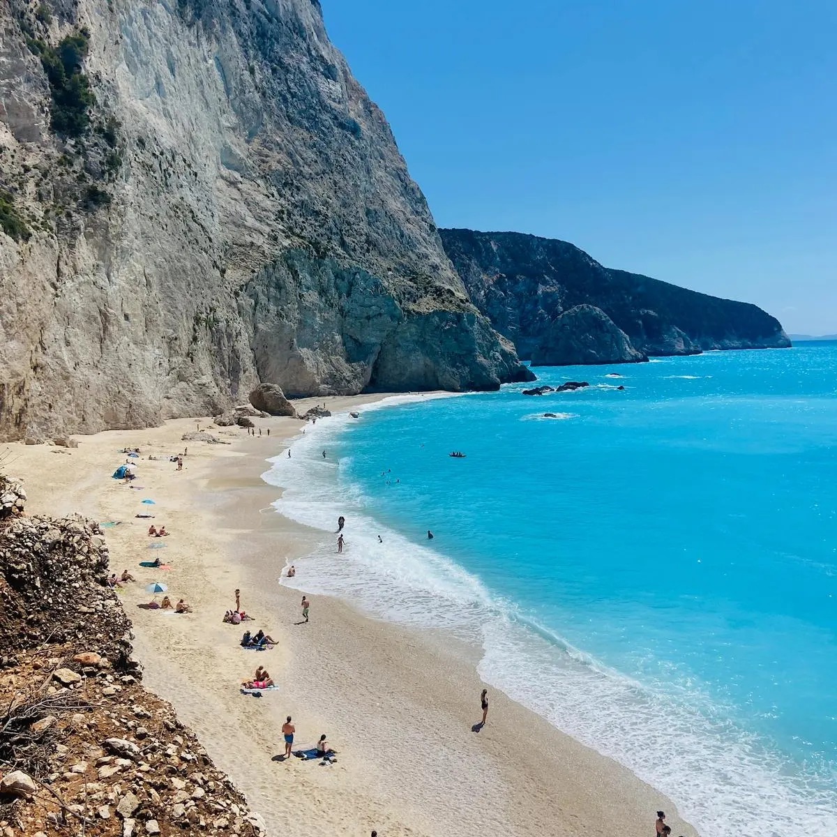 Turquoise beach with white sand between tall coastal cliffs, people relaxing on shore under blue sky
