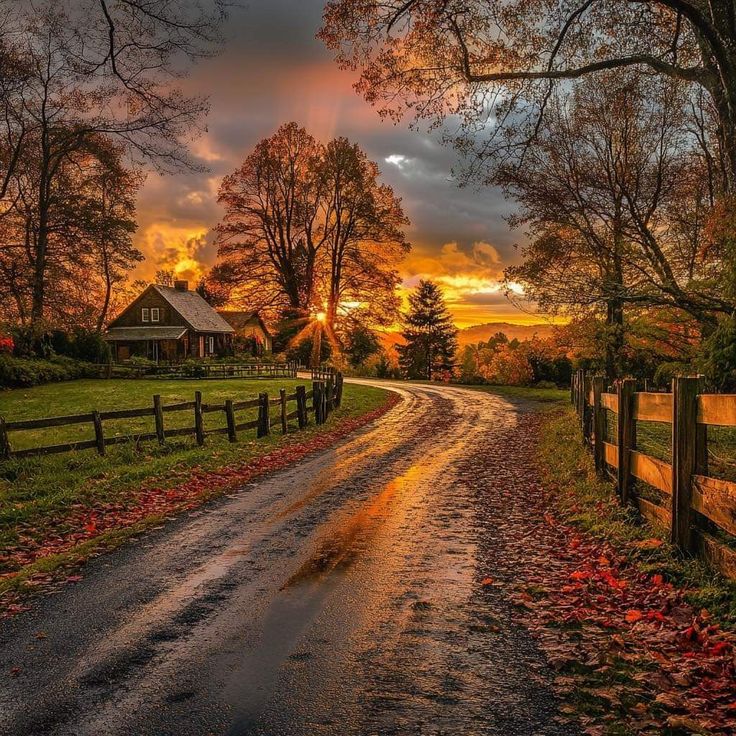 Country road at sunset with wooden fences, barn, and autumn trees under orange sky