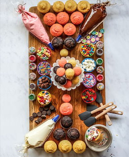 Wooden board with cupcakes, frosting ping bags, sprinkles, and colorful toppings arranged for decorating