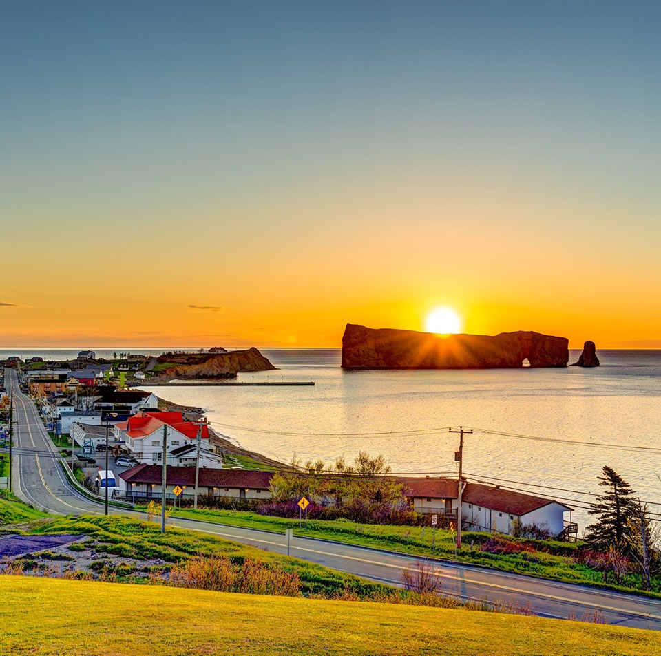 Sunset behind Percé Rock with calm water, coastal road, and houses in the foreground