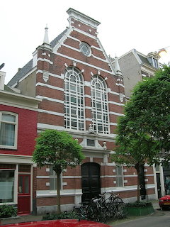 Red brick synagogue with large arched window and stepped gable on a tree-lined Amsterdam street