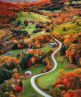 Winding road through a rural valley with colorful autumn trees, green fields, and scattered farmhouses