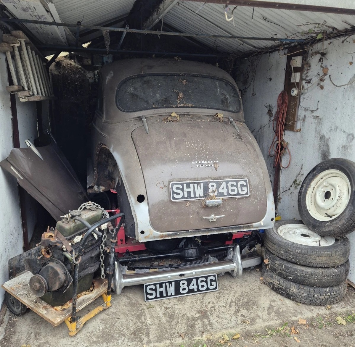 Old dusty Morris Minor car stored in a small garage with spare tires and an engine part
