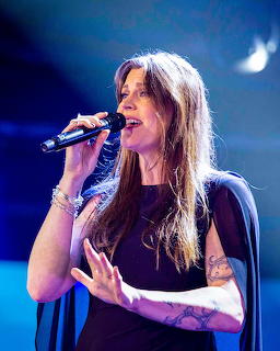 Woman with long brown hair singing into a microphone on stage under blue lighting