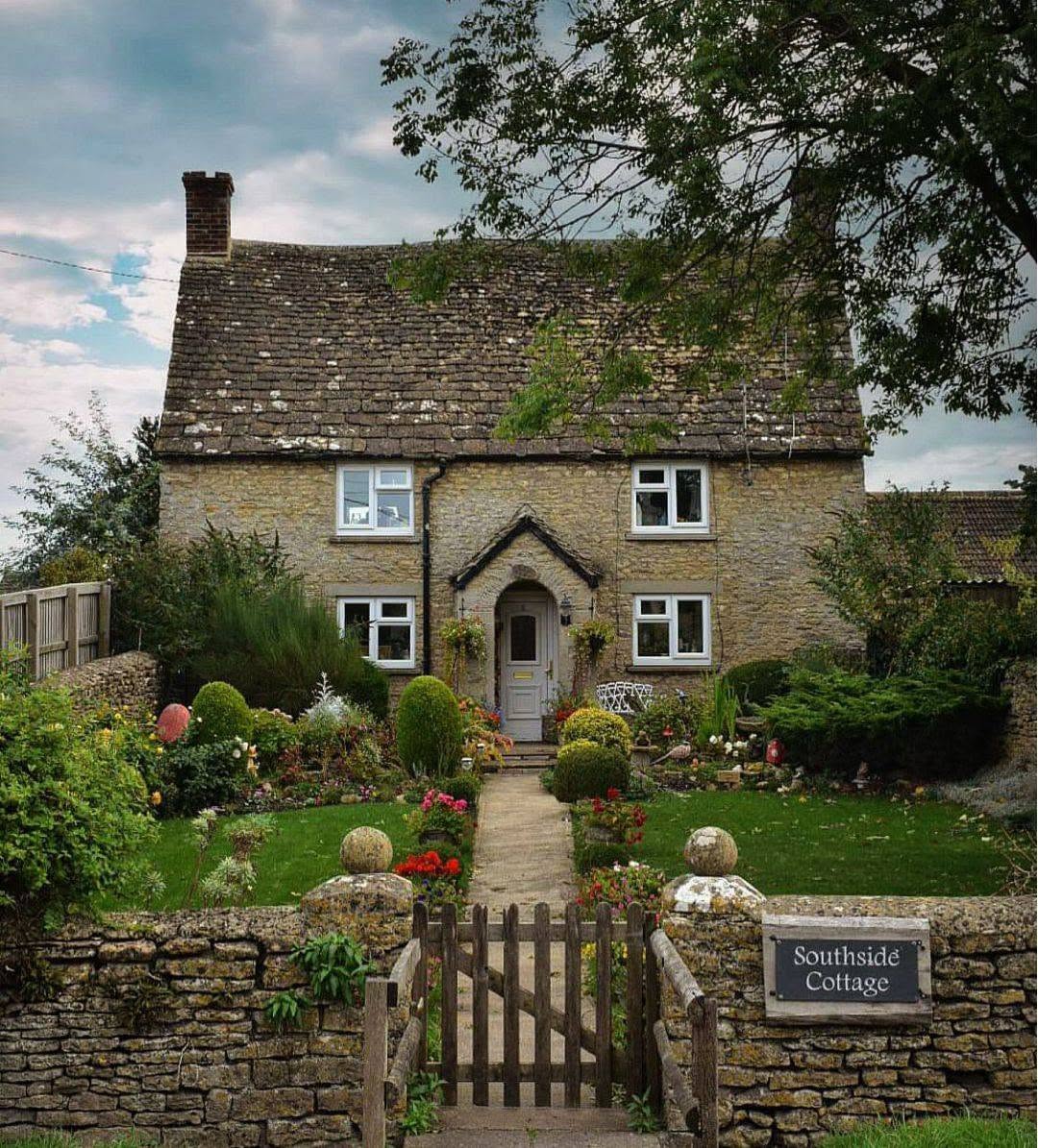 Stone cottage behind a wooden gate, garden, flowering shrubs, and low stone wall.