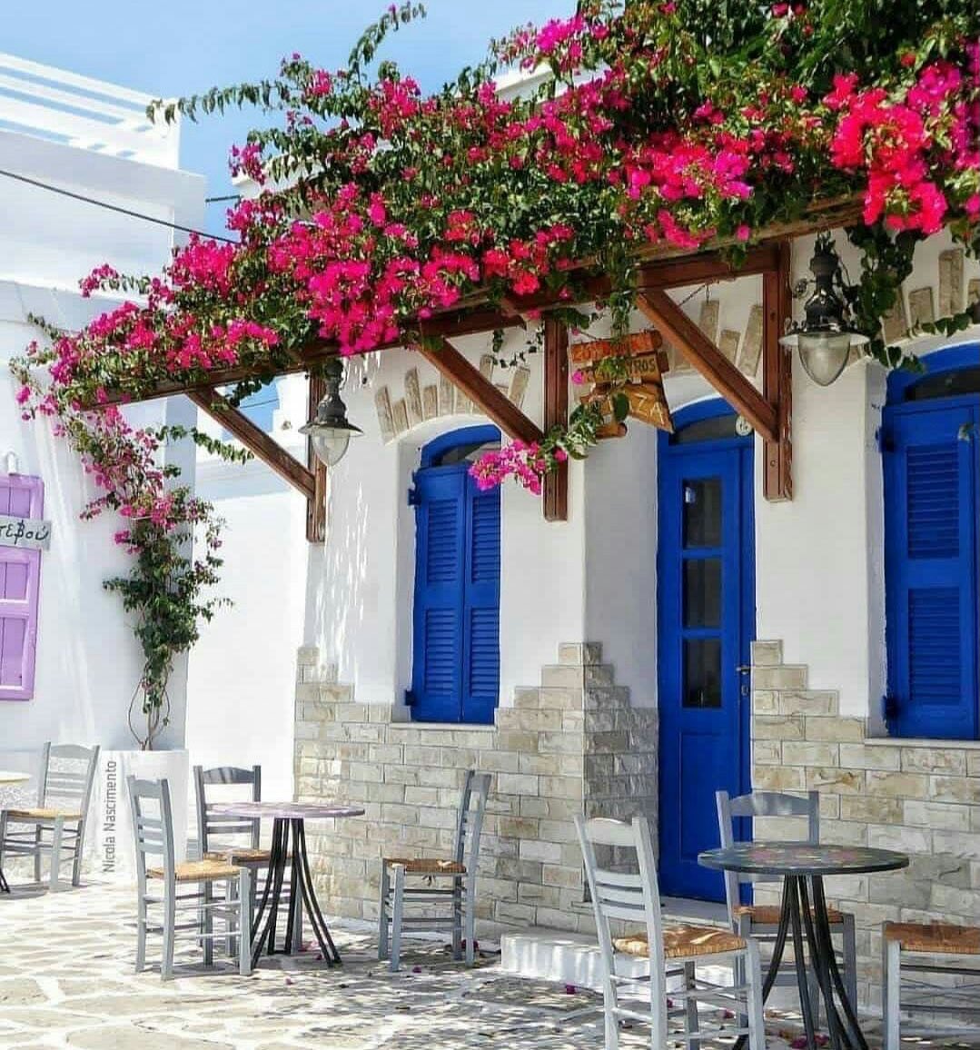 White Greek building with blue doors, pink bougainvillea overhead, white chairs on stone patio