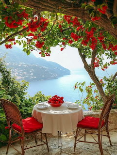 Terace with white tablecloth table and red chairs overlooking Mediterranean sea, framed by red flowers
