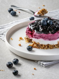 Slice of layered blueberry cheesecake with fresh blueberries and crumb crust on white plate