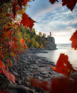 Lighthouse perched on rocky cliff beside calm water, framed by brilliant fall foliage in reds and oranges