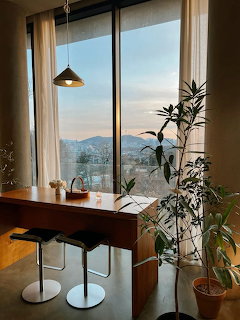 Cafe interior with wooden counter, bar stools, plants, and large window with mountain view at sunset