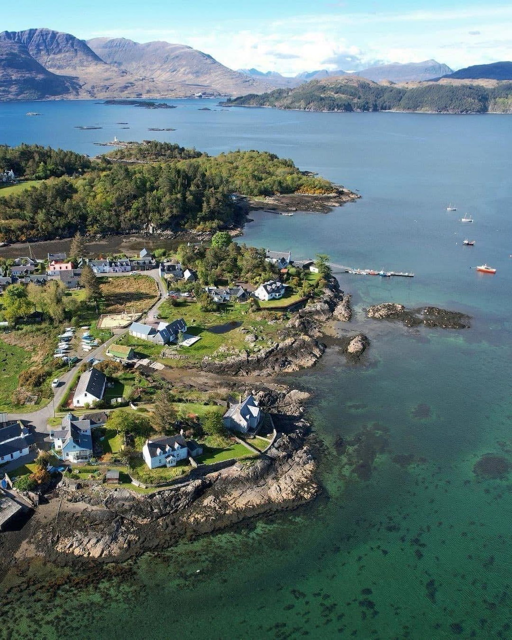 Aerial view of a small coastal Scottish village on a rocky shoreline with turquoise water and mountains beyond
