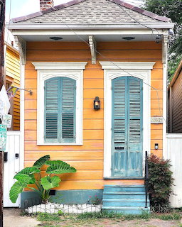 Small orange house with teal shutters and door, blue steps, and a large potted plant in front