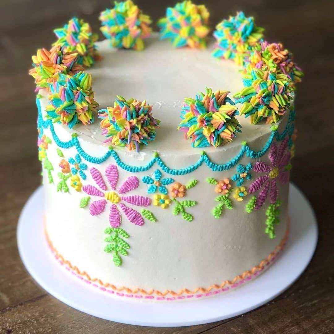 White frosted cake with colorful piped floral designs and rainbow pom-pom frosting decorations on top