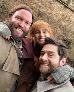 Three people in historical costumes smiling together in a selfie near a stone wall outdoors