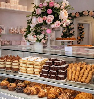 Bakery glass display case with rows of pastries, brownies, layered cakes, and croissants with pink flower arrangement above