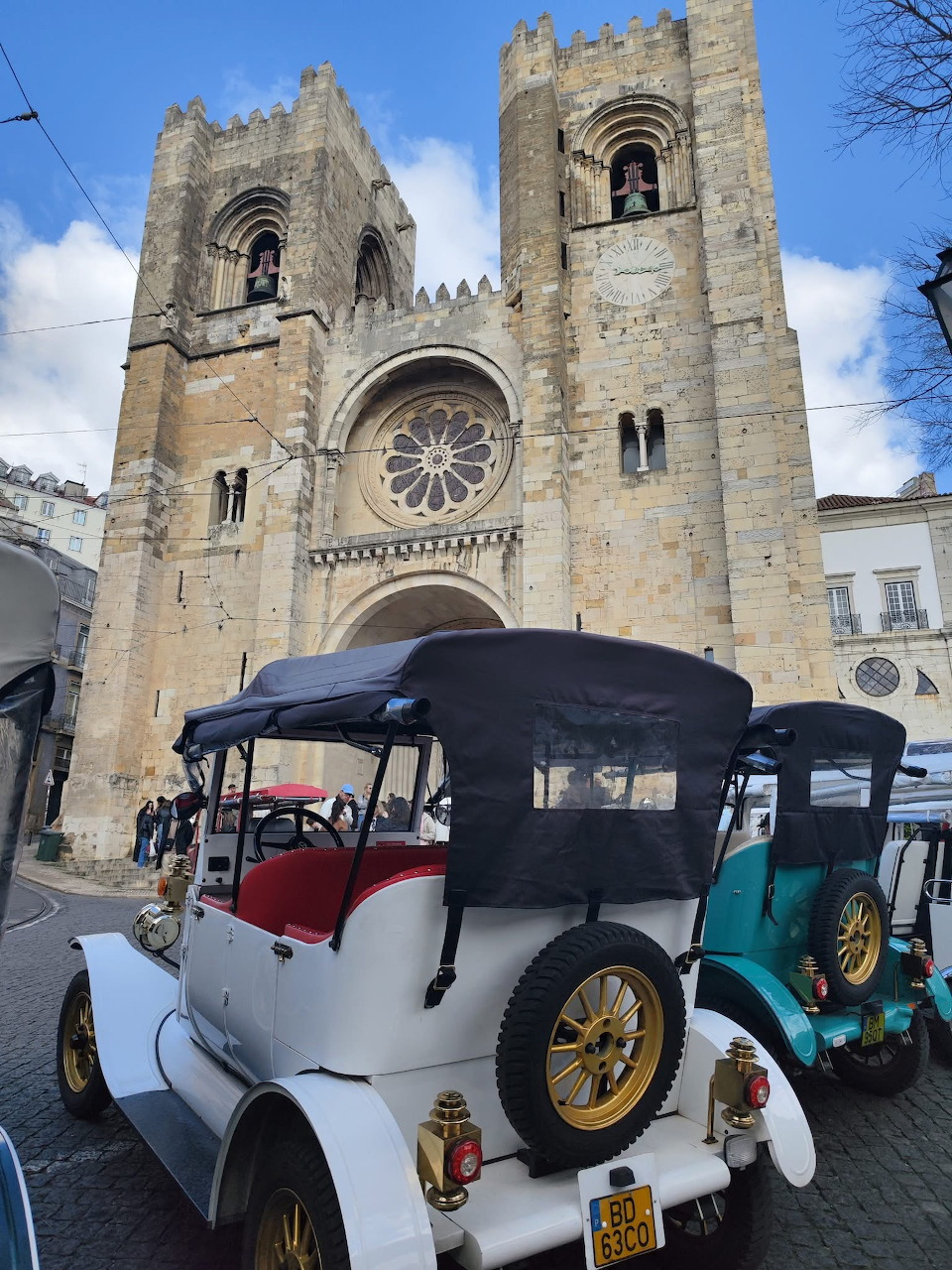 Lisbon cathedral with twin towers and rose window, vintage tuk-tuks parked in cobblestone square