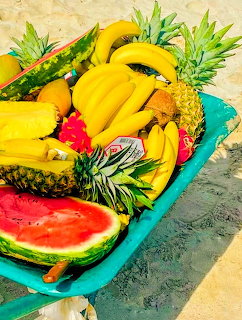 Turquoise cart overflowing with tropical fruits like bananas, pineapples, and watermelon on sand
