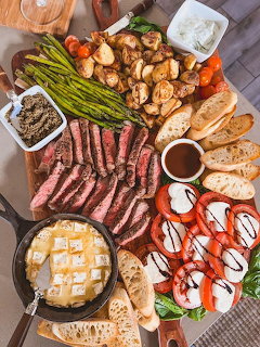 Overhead view of a meat and appetizer board with sliced steak, asparagus, caprese salad, bread, and dips