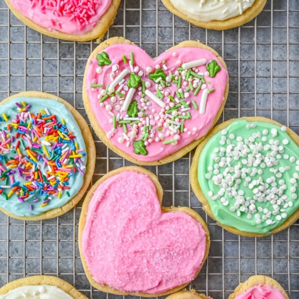 Heart-shaped and round sugar cookies with pink, green, and blue frosting decorated with colorful sprinkles on wire rack