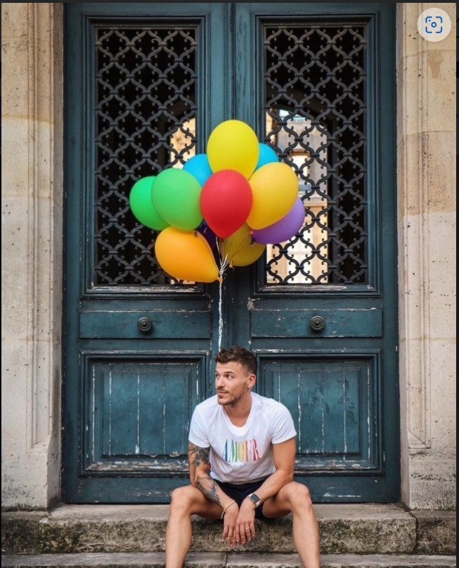 Young man sitting on steps holding colorful rainbow balloons in front of a teal ornate door