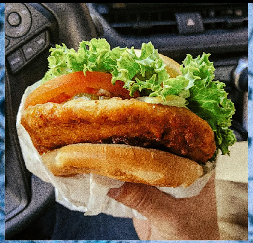 Hand holding a sausage sandwich with lettuce and tomato in a car