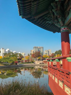 Korean pavilion with green roof and red pillars beside a reflecting pond with Seoul skyline and autumn trees