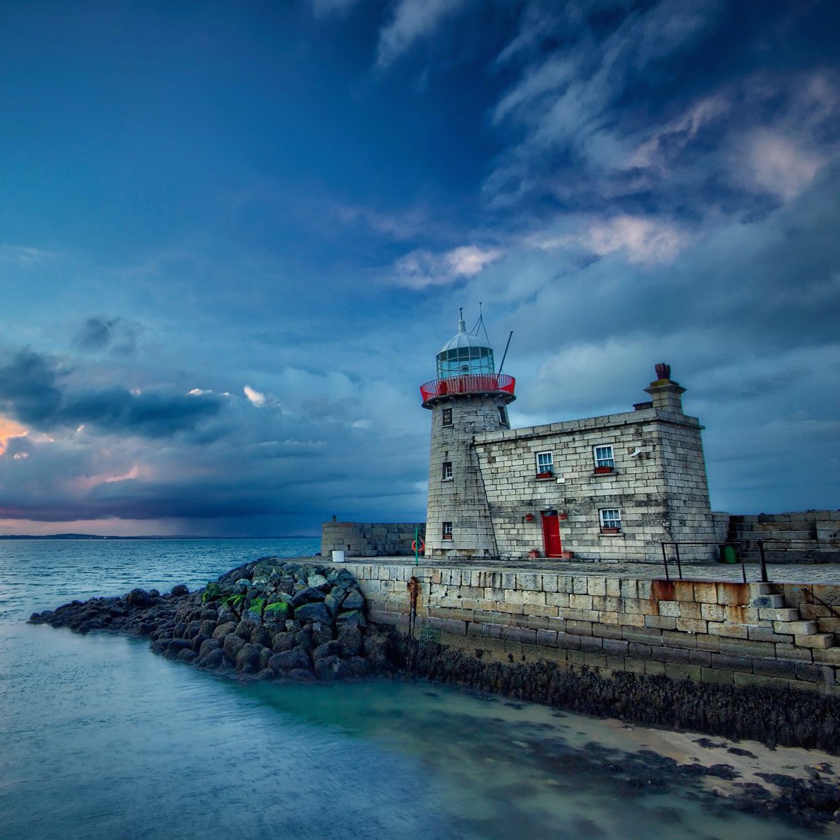 White lighthouse with red door on stone pier at dusk with dramatic cloudy sky and calm water