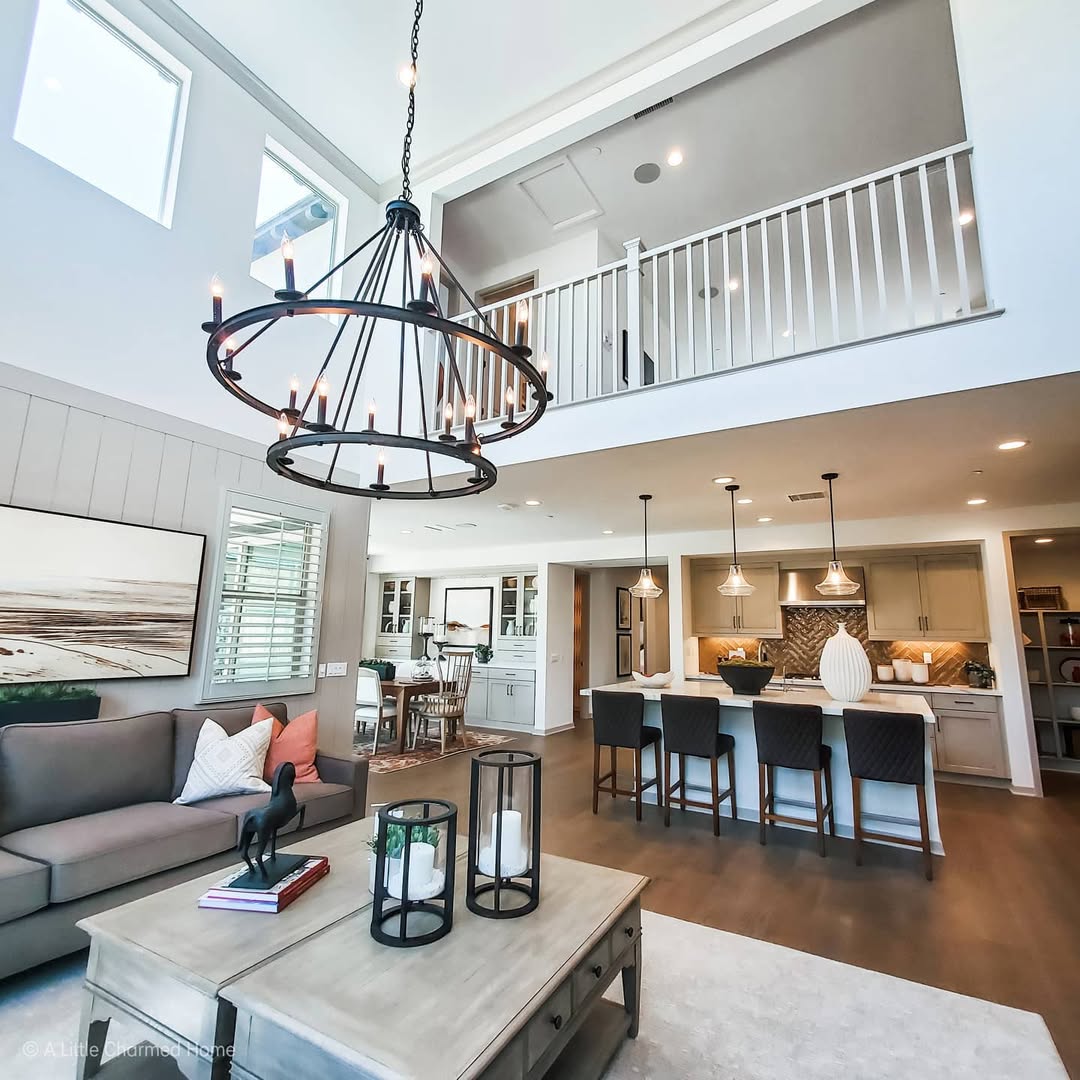 Bright open loft living room with chandelier, gray sofa, and kitchen island beneath an upstairs railing
