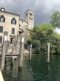Stone buildings and bell tower along a lake with wooden mooring posts and green water