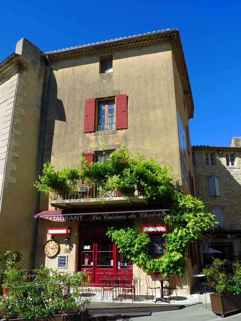 Stone building with red shutters and a restaurant facade covered in green vines in Gordes, France