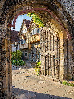 Medieval stone arch with carved columns framing Tudor half-timbered house and garden courtyard
