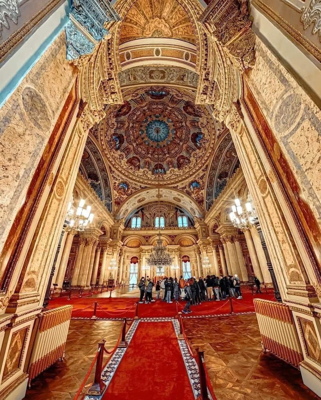 Ornate palace interior with decorated dome ceiling, chandeliers, red carpet and golden columns